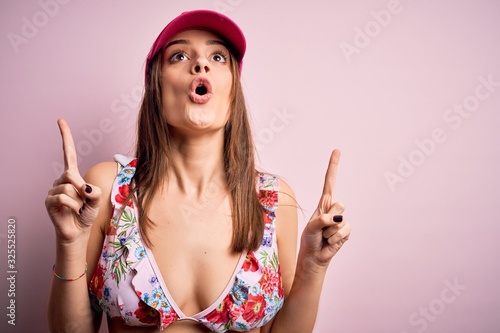 Young beautiful brunette woman on vacation wearing bikini and cap over pink background amazed and surprised looking up and pointing with fingers and raised arms.