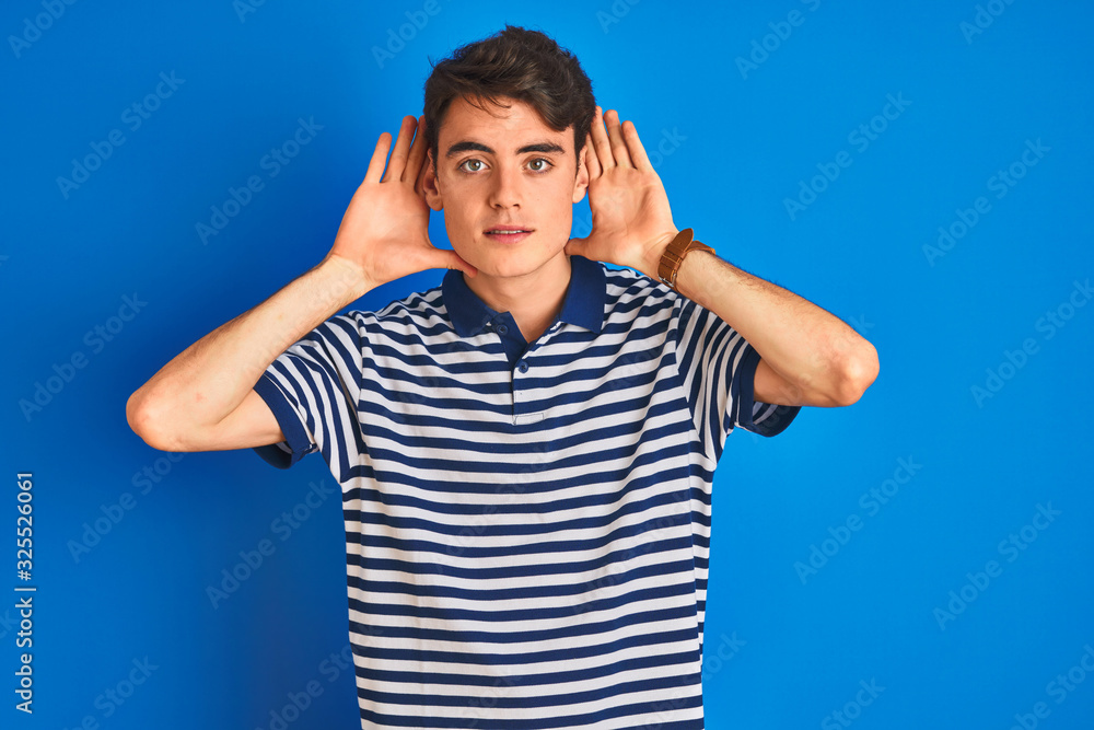 Teenager boy wearing casual t-shirt standing over blue isolated background Trying to hear both hands on ear gesture, curious for gossip. Hearing problem, deaf Teenager boy wearing casual t-shirt standing over blue isolated background Trying to hear both hands on ear gesture, curious for gossip. Hearing problem, deaf