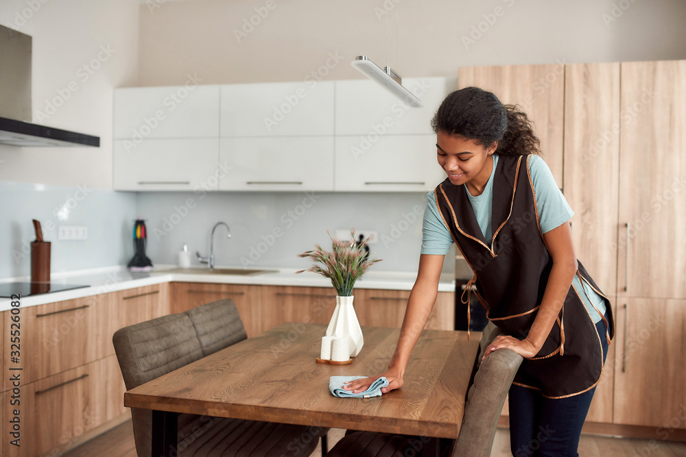 Pure clean. Young positive afro american house maid in apron cleaning table  in the modern kitchen. Professional female cleaner at work Stock Photo