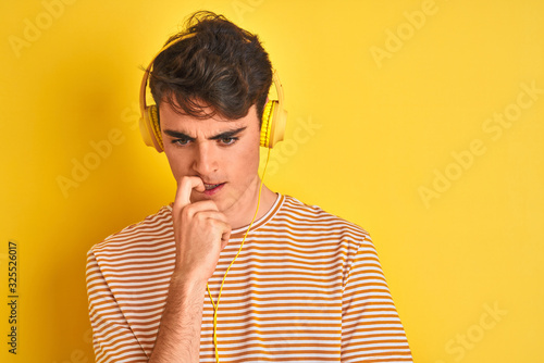 Teenager boy wearing headphones over isolated yellow background looking stressed and nervous with hands on mouth biting nails. Anxiety problem.