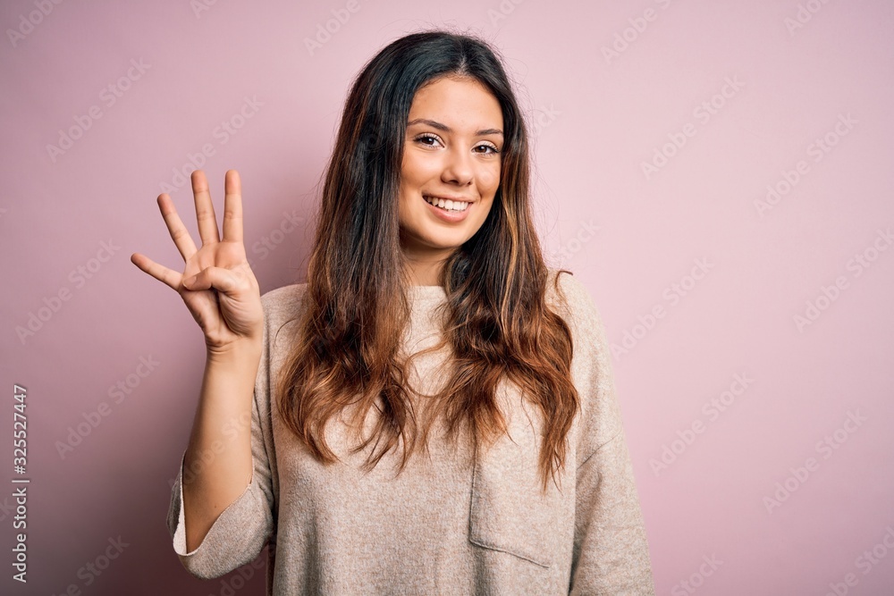 Young beautiful brunette woman wearing casual sweater standing over pink background showing and pointing up with fingers number four while smiling confident and happy. - obrazy, fototapety, plakaty 