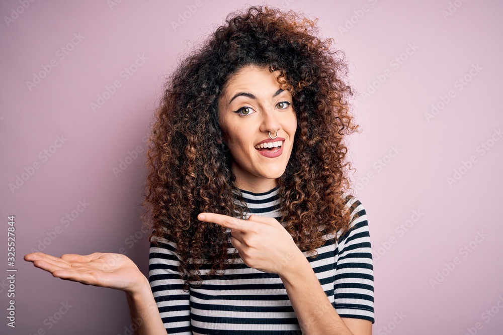 Young beautiful woman with curly hair and piercing wearing casual striped t-shirt amazed and smiling to the camera while presenting with hand and pointing with finger. Young beautiful woman with curly hair and piercing wearing casual striped t-shirt amazed and smiling to the camera while presenting with hand and pointing with finger.