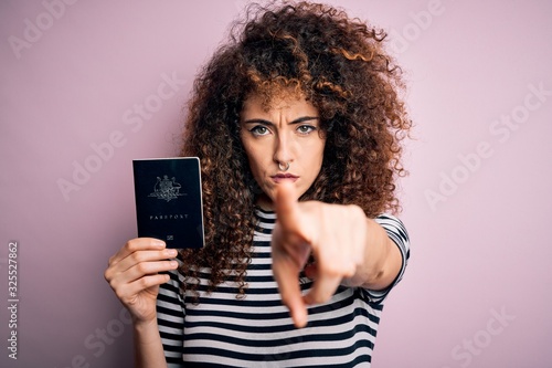 Beautiful tourist woman with curly hair and piercing holding australia australian passport id pointing with finger to the camera and to you, hand sign, positive and confident gesture from the front