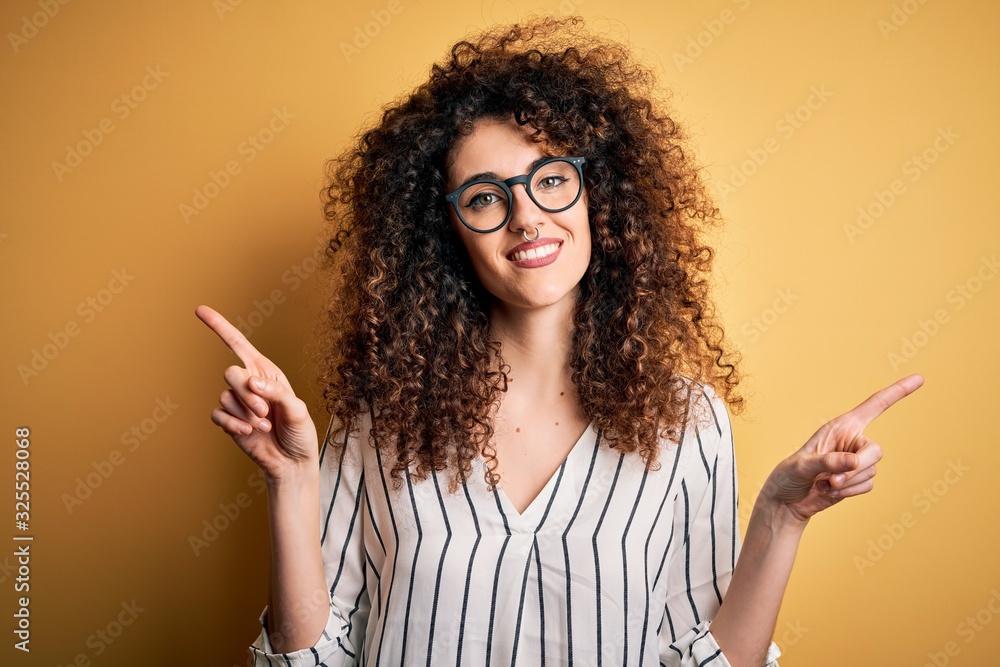 Young beautiful woman with curly hair and piercing wearing striped shirt and glasses smiling confident pointing with fingers to different directions. Copy space for advertisement Young beautiful woman with curly hair and piercing wearing striped shirt and glasses smiling confident pointing with fingers to different directions. Copy space for advertisement