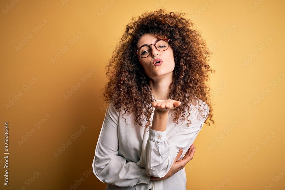 Young beautiful brunette woman with curly hair and piercing wearing shirt and glasses looking at the camera blowing a kiss with hand on air being lovely and sexy. Love expression. Young beautiful brunette woman with curly hair and piercing wearing shirt and glasses looking at the camera blowing a kiss with hand on air being lovely and sexy. Love expression.