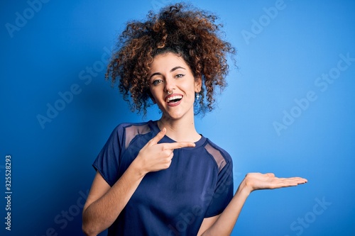 Young beautiful woman with curly hair and piercing wearing casual blue t-shirt amazed and smiling to the camera while presenting with hand and pointing with finger.