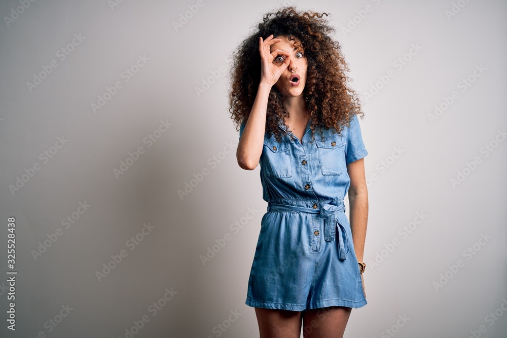 Young beautiful woman with curly hair and piercing wearing casual denim dress doing ok gesture shocked with surprised face, eye looking through fingers. Unbelieving expression. Young beautiful woman with curly hair and piercing wearing casual denim dress doing ok gesture shocked with surprised face, eye looking through fingers. Unbelieving expression.