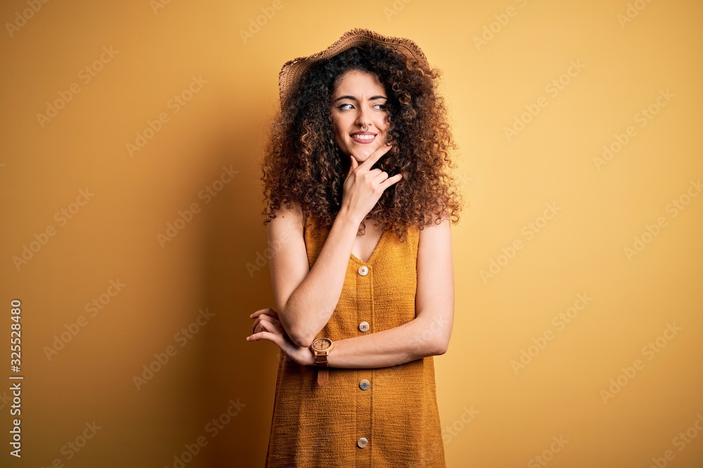 Beautiful brunette woman on vacation with curly hair and piercing wearing hat and dress with hand on chin thinking about question, pensive expression. Smiling with thoughtful face. Doubt concept. Beautiful brunette woman on vacation with curly hair and piercing wearing hat and dress with hand on chin thinking about question, pensive expression. Smiling with thoughtful face. Doubt concept.