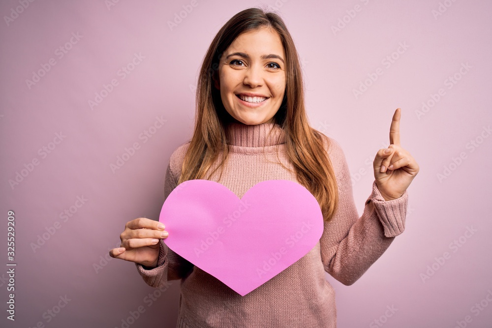 Young blonde woman holding romantic big paper heart shape over pink isolated background surprised with an idea or question pointing finger with happy face, number one Young blonde woman holding romantic big paper heart shape over pink isolated background surprised with an idea or question pointing finger with happy face, number one