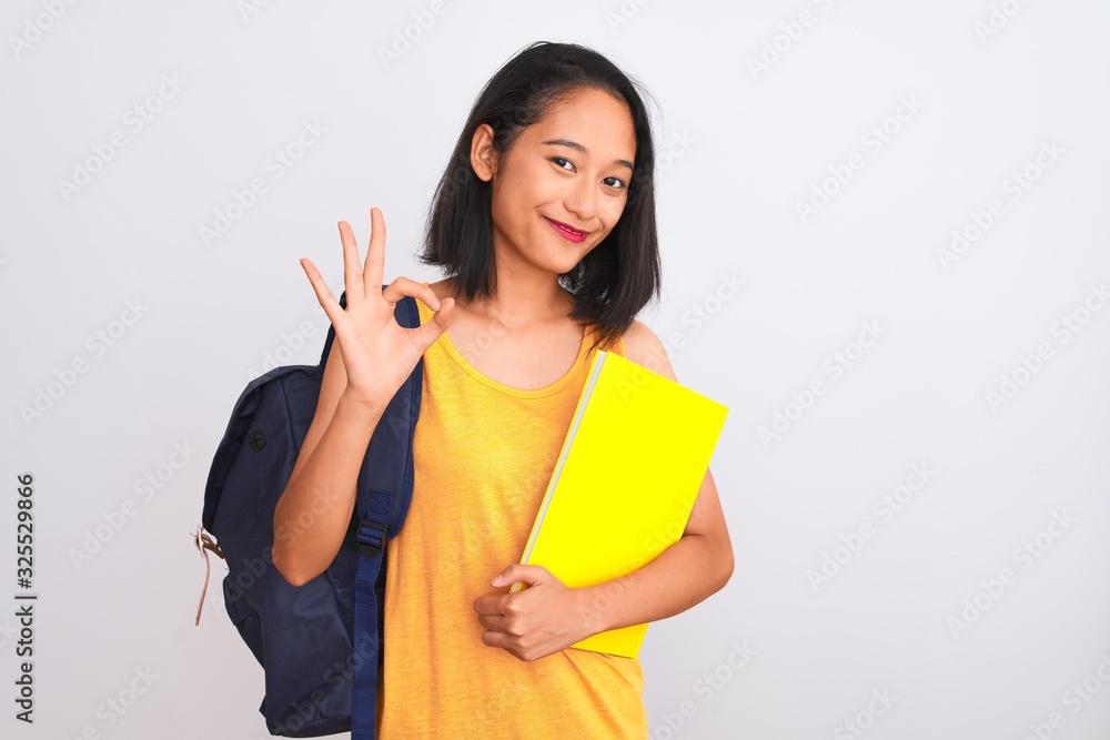 Young chinese student woman wearing backpack holding book over isolated white background doing ok sign with fingers, excellent symbol Young chinese student woman wearing backpack holding book over isolated white background doing ok sign with fingers, excellent symbol