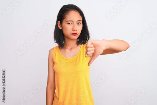 Young chinese woman wearing yellow casual t-shirt standing over isolated white background looking unhappy and angry showing rejection and negative with thumbs down gesture. Bad expression.