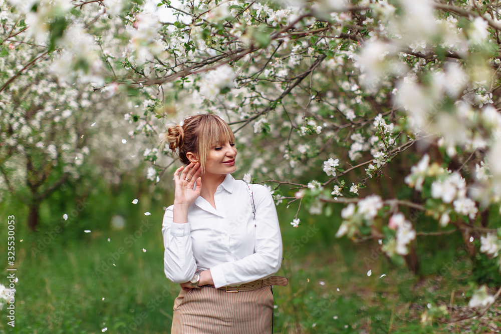 Beautiful young blonde woman in white shirt posing under apple tree in blossom in Spring garden Beautiful young blonde woman in white shirt posing under apple tree in blossom in Spring garden
