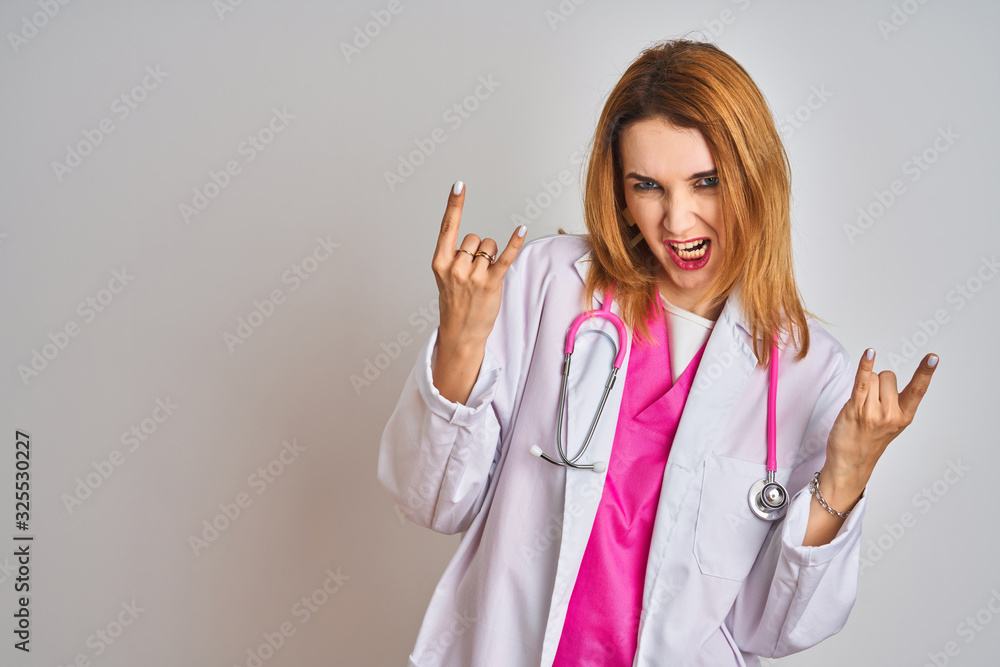 Redhead caucasian doctor woman wearing pink stethoscope over isolated background shouting with crazy expression doing rock symbol with hands up. Music star. Heavy concept.