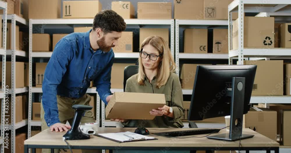 Caucasian woman postal worker working at computer in office store. Postman bringing carton box and female colleague redistering it while couple talking. Mail delivery registration. Caucasian woman postal worker working at computer in office store. Postman bringing carton box and female colleague redistering it while couple talking. Mail delivery registration.