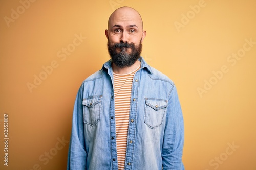 Handsome bald man with beard wearing casual denim jacket and striped t-shirt puffing cheeks with funny face. Mouth inflated with air, crazy expression.