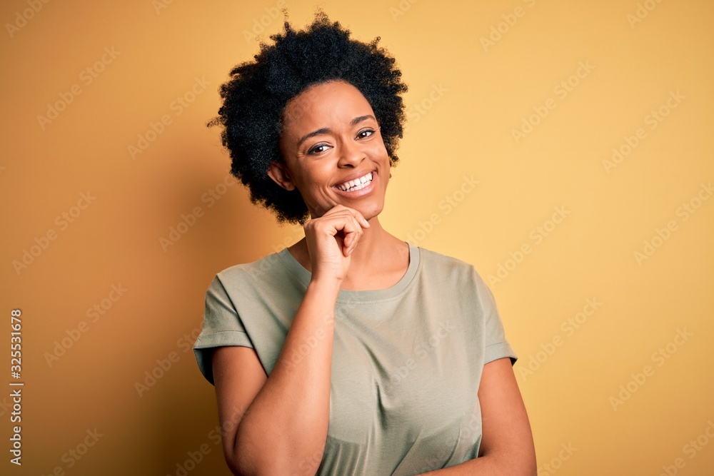 Young beautiful African American afro woman with curly hair wearing casual t-shirt looking confident at the camera with smile with crossed arms and hand raised on chin. Thinking positive. Young beautiful African American afro woman with curly hair wearing casual t-shirt looking confident at the camera with smile with crossed arms and hand raised on chin. Thinking positive.