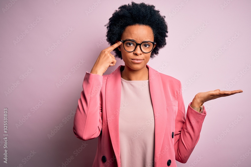 Young beautiful African American afro businesswoman with curly hair wearing pink jacket confused and annoyed with open palm showing copy space and pointing finger to forehead. Think about it. Young beautiful African American afro businesswoman with curly hair wearing pink jacket confused and annoyed with open palm showing copy space and pointing finger to forehead. Think about it.