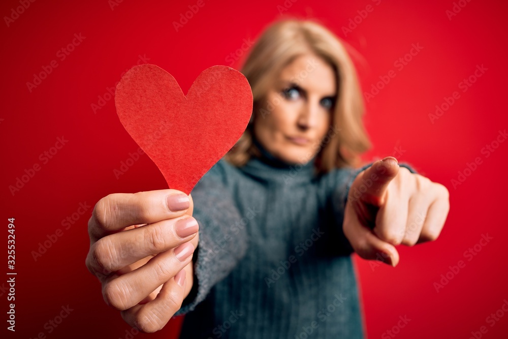 Middle age beautiful blonde romantic woman holding red paper heart celebrating valentine day pointing with finger to the camera and to you, hand sign, positive and confident gesture from the front Middle age beautiful blonde romantic woman holding red paper heart celebrating valentine day pointing with finger to the camera and to you, hand sign, positive and confident gesture from the front
