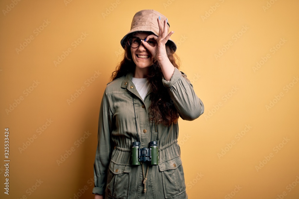 Young beautiful tourist woman on vacation wearing explorer hat and binoculars doing ok gesture with hand smiling, eye looking through fingers with happy face. Young beautiful tourist woman on vacation wearing explorer hat and binoculars doing ok gesture with hand smiling, eye looking through fingers with happy face.
