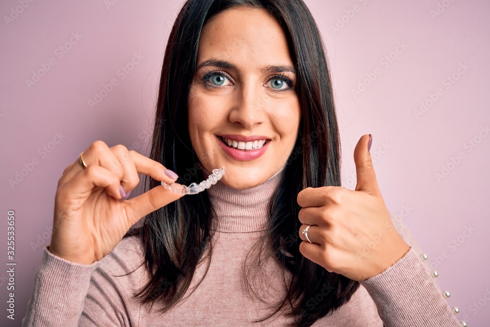 Young woman with blue eyes holding clear aligner standing over pink background happy with big smile doing ok sign, thumb up with fingers, excellent sign Young woman with blue eyes holding clear aligner standing over pink background happy with big smile doing ok sign, thumb up with fingers, excellent sign