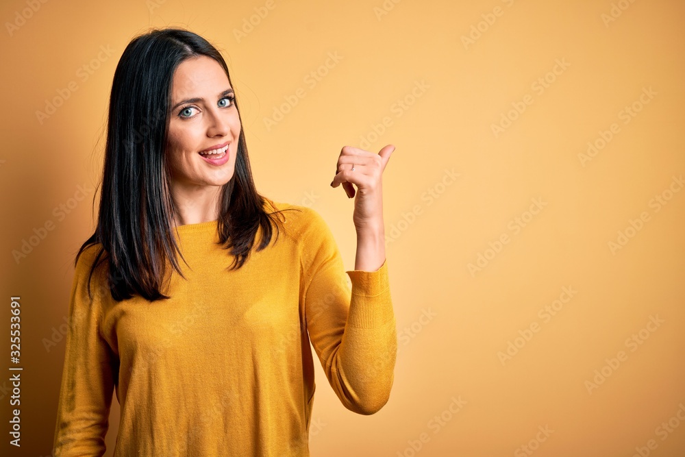 Young brunette woman with blue eyes wearing casual sweater over yellow background smiling with happy face looking and pointing to the side with thumb up. Young brunette woman with blue eyes wearing casual sweater over yellow background smiling with happy face looking and pointing to the side with thumb up.
