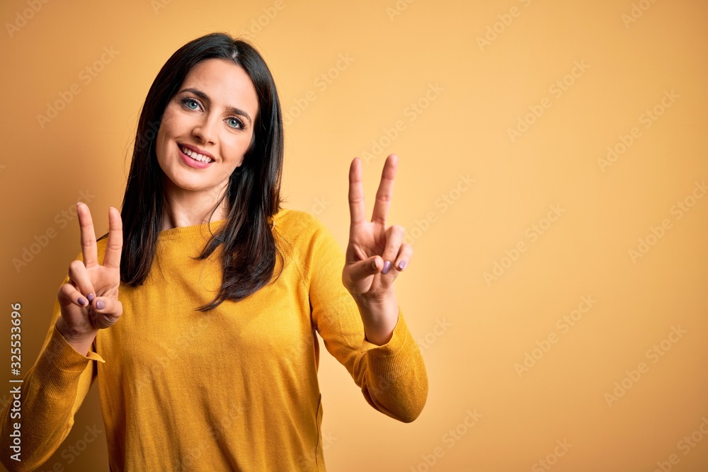 Young brunette woman with blue eyes wearing casual sweater over yellow background smiling looking to the camera showing fingers doing victory sign. Number two.