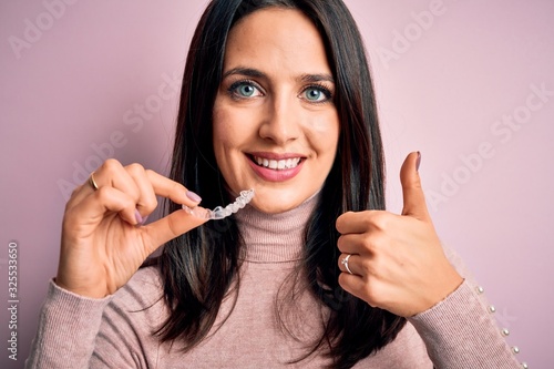 Young woman with blue eyes holding clear aligner standing over pink background happy with big smile doing ok sign, thumb up with fingers, excellent sign
