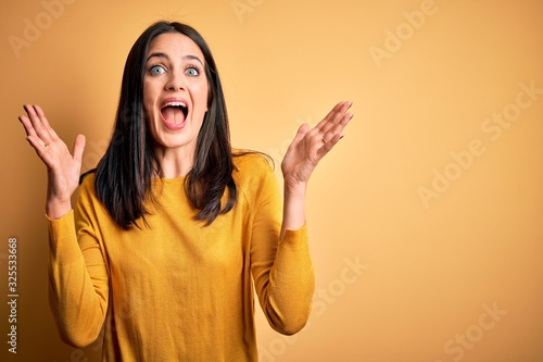 Young brunette woman with blue eyes wearing casual sweater over yellow background celebrating crazy and amazed for success with arms raised and open eyes screaming excited. Winner concept