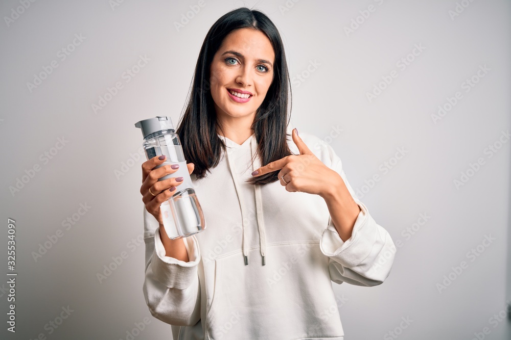 Young brunette sporty woman with blue eyes holding water bottle over white background with surprise face pointing finger to himself Young brunette sporty woman with blue eyes holding water bottle over white background with surprise face pointing finger to himself