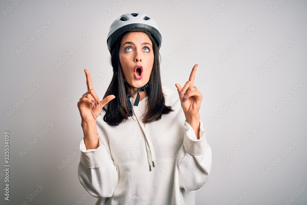 Young cyclist woman with blue eyes wearing bike helmet over isolated white background amazed and surprised looking up and pointing with fingers and raised arms. Young cyclist woman with blue eyes wearing bike helmet over isolated white background amazed and surprised looking up and pointing with fingers and raised arms.