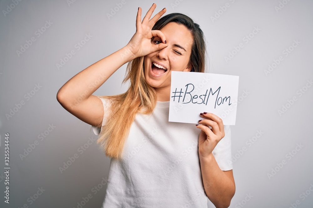 Young beautiful woman holding paper with best mom message celebrating mothers day with happy face smiling doing ok sign with hand on eye looking through fingers Young beautiful woman holding paper with best mom message celebrating mothers day with happy face smiling doing ok sign with hand on eye looking through fingers
