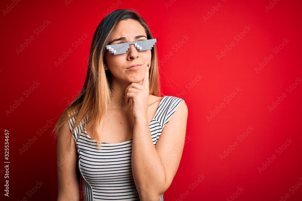 Young beautiful brunette woman wearing funny thug life sunglasses over red background with hand on chin thinking about question, pensive expression. Smiling with thoughtful face. Doubt concept.