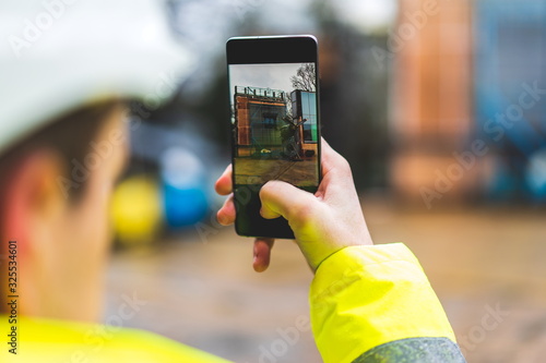 Architect holding a smartphone on construction site - young construction worker is using mobile phone on site - Construction worker with building plans and cellphone - Focus on mobile. warm filter