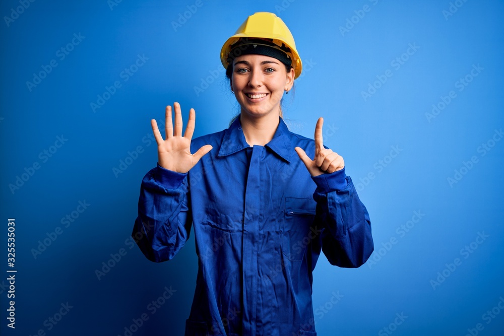 Young beautiful worker woman with blue eyes wearing security helmet and uniform showing and pointing up with fingers number seven while smiling confident and happy.