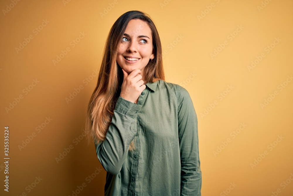 Young beautiful blonde woman with blue eyes wearing green shirt over yellow background with hand on chin thinking about question, pensive expression. Smiling with thoughtful face. Doubt concept. Young beautiful blonde woman with blue eyes wearing green shirt over yellow background with hand on chin thinking about question, pensive expression. Smiling with thoughtful face. Doubt concept.