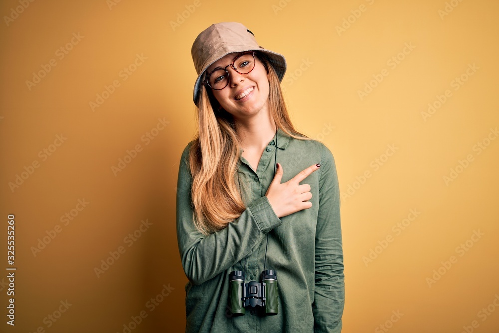 Beautiful blonde explorer woman with blue eyes wearing hat and glasses using binoculars cheerful with a smile of face pointing with hand and finger up to the side with happy and natural expression Beautiful blonde explorer woman with blue eyes wearing hat and glasses using binoculars cheerful with a smile of face pointing with hand and finger up to the side with happy and natural expression
