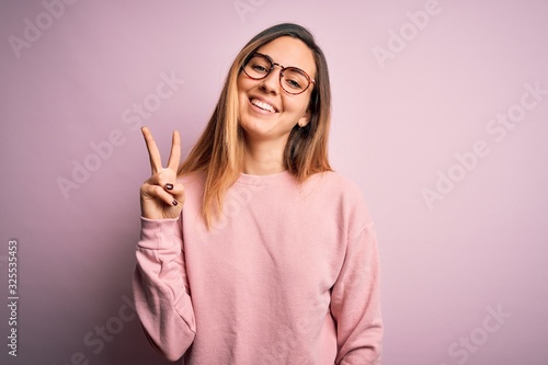 Beautiful blonde woman with blue eyes wearing sweater and glasses over pink background showing and pointing up with fingers number two while smiling confident and happy. © Krakenimages.com