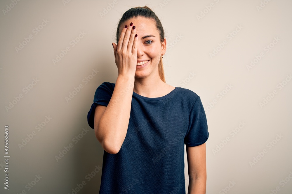 Young beautiful blonde woman with blue eyes wearing casual t-shirt over white background covering one eye with hand, confident smile on face and surprise emotion. Young beautiful blonde woman with blue eyes wearing casual t-shirt over white background covering one eye with hand, confident smile on face and surprise emotion.