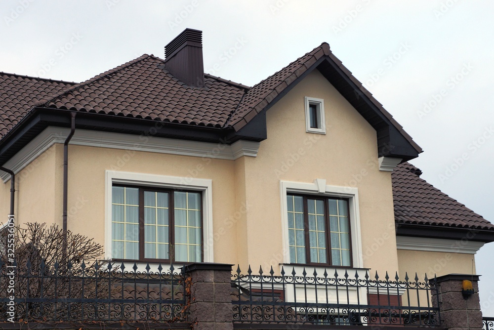 brown private house with windows under a tiled roof against a gray sky behind a fence 