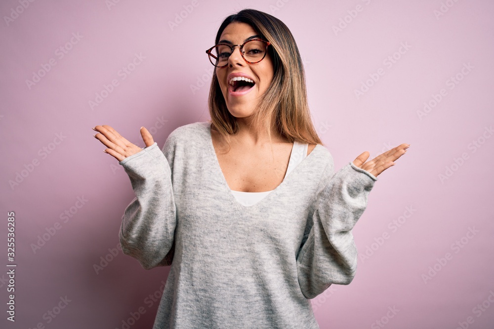 Young beautiful brunette woman wearing casual sweater and glasses over pink background celebrating crazy and amazed for success with arms raised and open eyes screaming excited. Winner concept