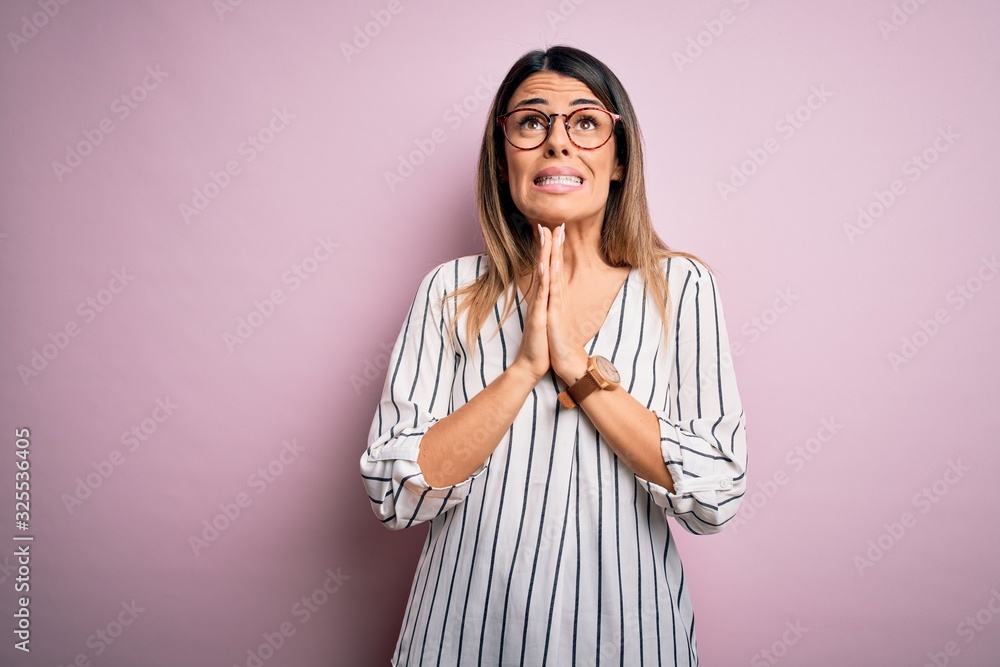 Young beautiful woman wearing casual striped t-shirt and glasses over pink background begging and praying with hands together with hope expression on face very emotional and worried. Begging. Young beautiful woman wearing casual striped t-shirt and glasses over pink background begging and praying with hands together with hope expression on face very emotional and worried. Begging.