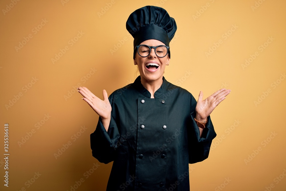 Young beautiful brunette chef woman wearing cooker uniform and hat over yellow background celebrating mad and crazy for success with arms raised and closed eyes screaming excited. Winner concept Young beautiful brunette chef woman wearing cooker uniform and hat over yellow background celebrating mad and crazy for success with arms raised and closed eyes screaming excited. Winner concept