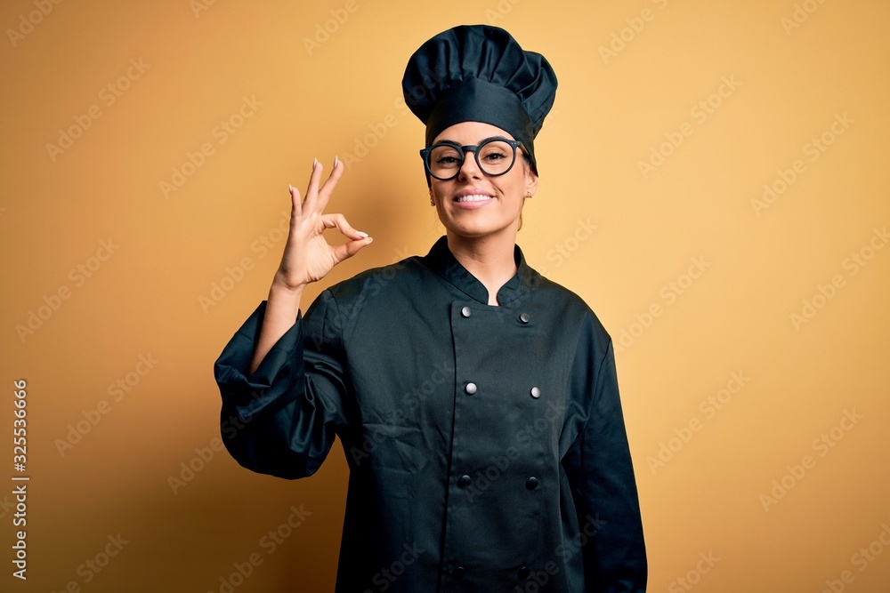 Young beautiful brunette chef woman wearing cooker uniform and hat over yellow background smiling positive doing ok sign with hand and fingers. Successful expression. Young beautiful brunette chef woman wearing cooker uniform and hat over yellow background smiling positive doing ok sign with hand and fingers. Successful expression.
