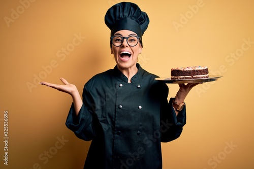 Young beautiful brunette baker woman wearing cooker uniform and hat holding cake very happy and excited, winner expression celebrating victory screaming with big smile and raised hands photo