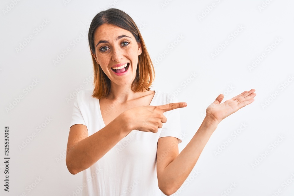 Beautiful redhead woman wearing casual white t-shirt over isolated background amazed and smiling to the camera while presenting with hand and pointing with finger. Beautiful redhead woman wearing casual white t-shirt over isolated background amazed and smiling to the camera while presenting with hand and pointing with finger.
