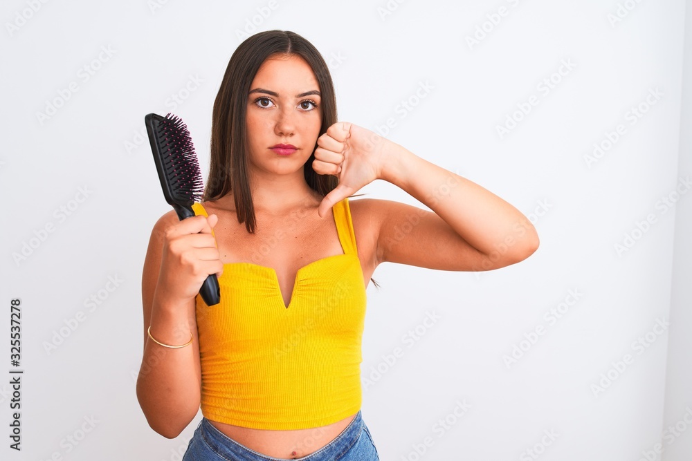 Young beautiful girl holding hair comb standing over isolated white background with angry face, negative sign showing dislike with thumbs down, rejection concept Young beautiful girl holding hair comb standing over isolated white background with angry face, negative sign showing dislike with thumbs down, rejection concept