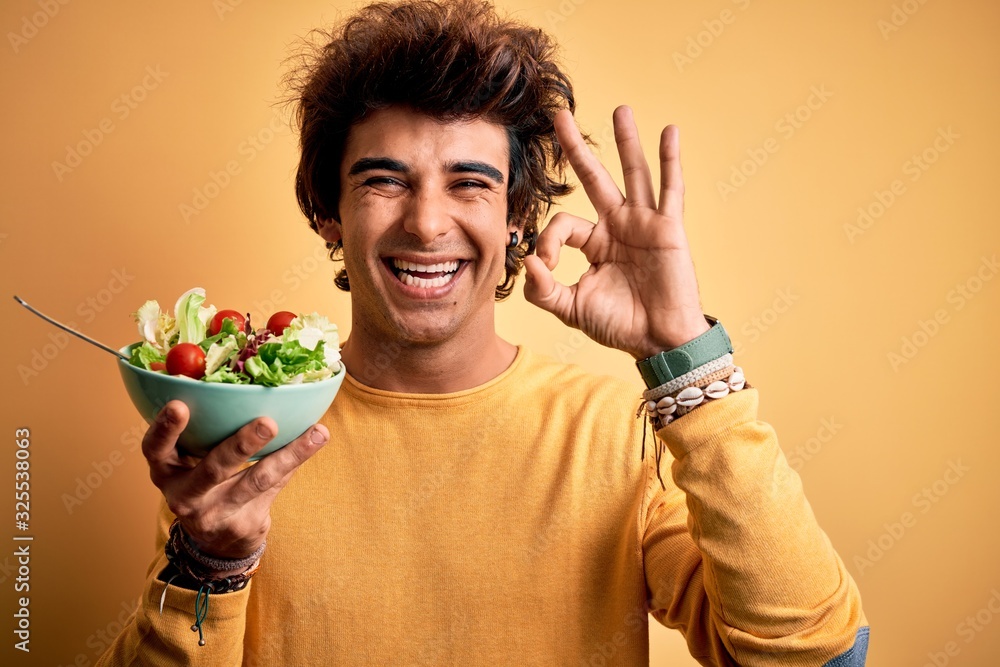 Young handsome man holding bowl with salad standing over isolated yellow background doing ok sign with fingers, excellent symbol Young handsome man holding bowl with salad standing over isolated yellow background doing ok sign with fingers, excellent symbol
