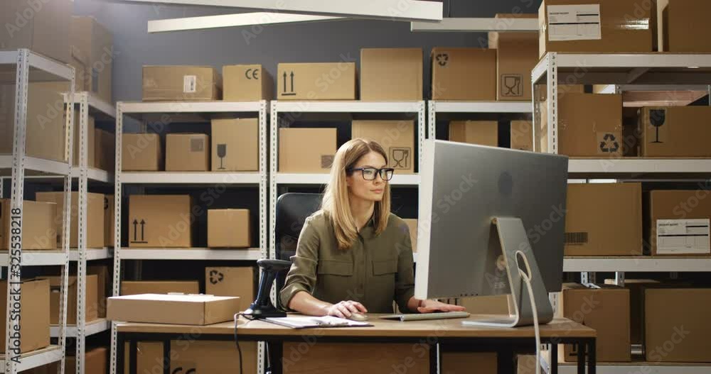 Caucasian beautiful female postal worker working at computer in post delivery office and typing on keyboard. Young woman in mail store sitting at table in storage. Caucasian beautiful female postal worker working at computer in post delivery office and typing on keyboard. Young woman in mail store sitting at table in storage.