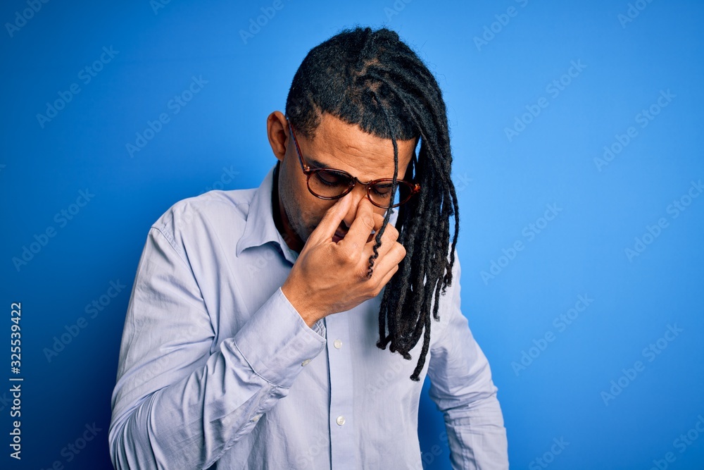 Young handsome african american man with dreadlocks wearing casual shirt and glasses tired rubbing nose and eyes feeling fatigue and headache. Stress and frustration concept. Young handsome african american man with dreadlocks wearing casual shirt and glasses tired rubbing nose and eyes feeling fatigue and headache. Stress and frustration concept.