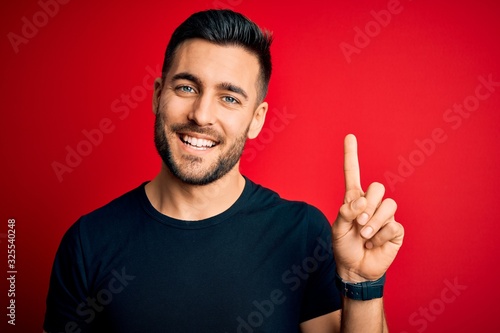 Young handsome man wearing casual black t-shirt standing over isolated red background showing and pointing up with finger number one while smiling confident and happy.